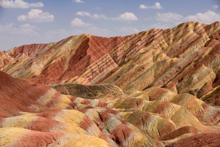 Le nord de la Chine s’est ouvert ces dernières années au tourisme, permettant de découvrir différentes régions aux paysages exceptionnels. Dans la province de Gansu, le Parc géologique national (géoparc) de Zhangye Danxia, créé en 2011, est une vaste zone de 500 km2 de formations géologiques colorées composées de grès et d’argiles. Ce site, dont le nom danxia signifie en chinois « relief de nuages rouges », est inscrit au patrimoine mondial de l’UNESCO depuis 2010. Il est situé à une trentaine de kilomètres à l’ouest de la ville de Zanghie. Il se compose d’une succession de collines aux reliefs érodés présentant des couches dont les couleurs varient du gris bleuté au rouge en passant par toutes les nuances de jaune, orange, et rose. Alors que ce site était encore isolé et méconnu il y a environ cinq ans, a surgi depuis peu en limite du parc une agglomération composée essentiellement d’hôtels, ce qui laisse augurer d’un développement touristique très rapide dans les prochaines années. L’accès au parc est très règlementé : une navette de bus conduit les visiteurs aux principaux belvédères, au nombre de cinq (mais dont seulement trois étaient ouverts au public en avril 2016). Une série d’escaliers permettent ainsi d’accéder à des plateformes offrant une vue panoramique sur le paysage. En contrepartie, il est impossible de marcher en dehors des endroits aménagés, pas même sur la route où seuls les bus ont le droit de circuler. C’est évidemment un paradis pour photographes, surtout les jours de soleil, car les toutes les nuances sont visibles et contrastent avec le bleu du ciel. Le belvédère n°1, proche de l’entrée nord, permet d’avoir une vue d’ensemble sur plusieurs paysages très colorés, alors que le belvédère n°4 domine une des scènes emblématiques du site : des couches géologiques inclinées formant de fines rayures multicolores. Même si certaines photos publiées sur Internet exagèrent délibérément les couleurs (merci Photoshop), il n’en reste pas moins que Danxia constitue un paysage naturel tout à fait exceptionnel.