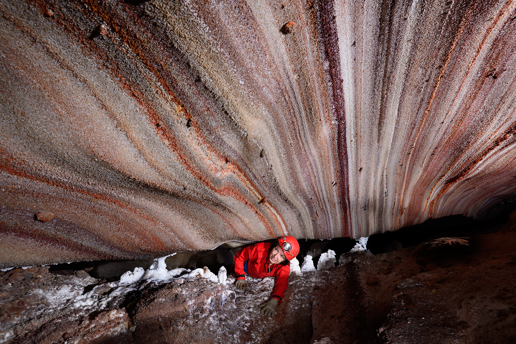 3N Cave (Iran, île de Qeshm, diapir de sel de Namakdan) - Entrée supérieure : spéléo passant une étroiture avec des couches de sel colorées au plafond (blanc, rose, orangé, rouge)