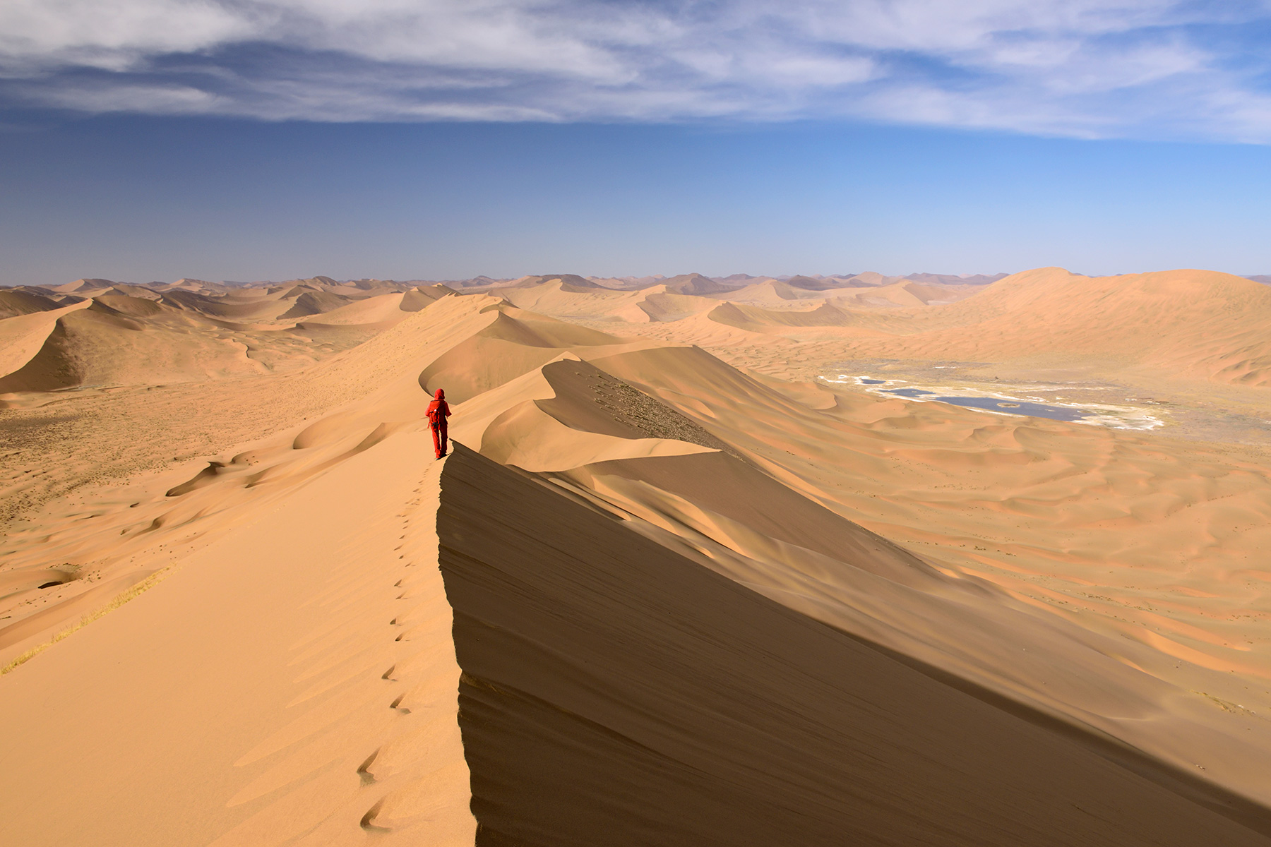 Désert du Badain Jaran (Chine, Mongolie Intérieure) - Vue générale d'un enchaînement de grandes dunes avec randonneuse sur la crête. Vallée avec lac salé à droite.