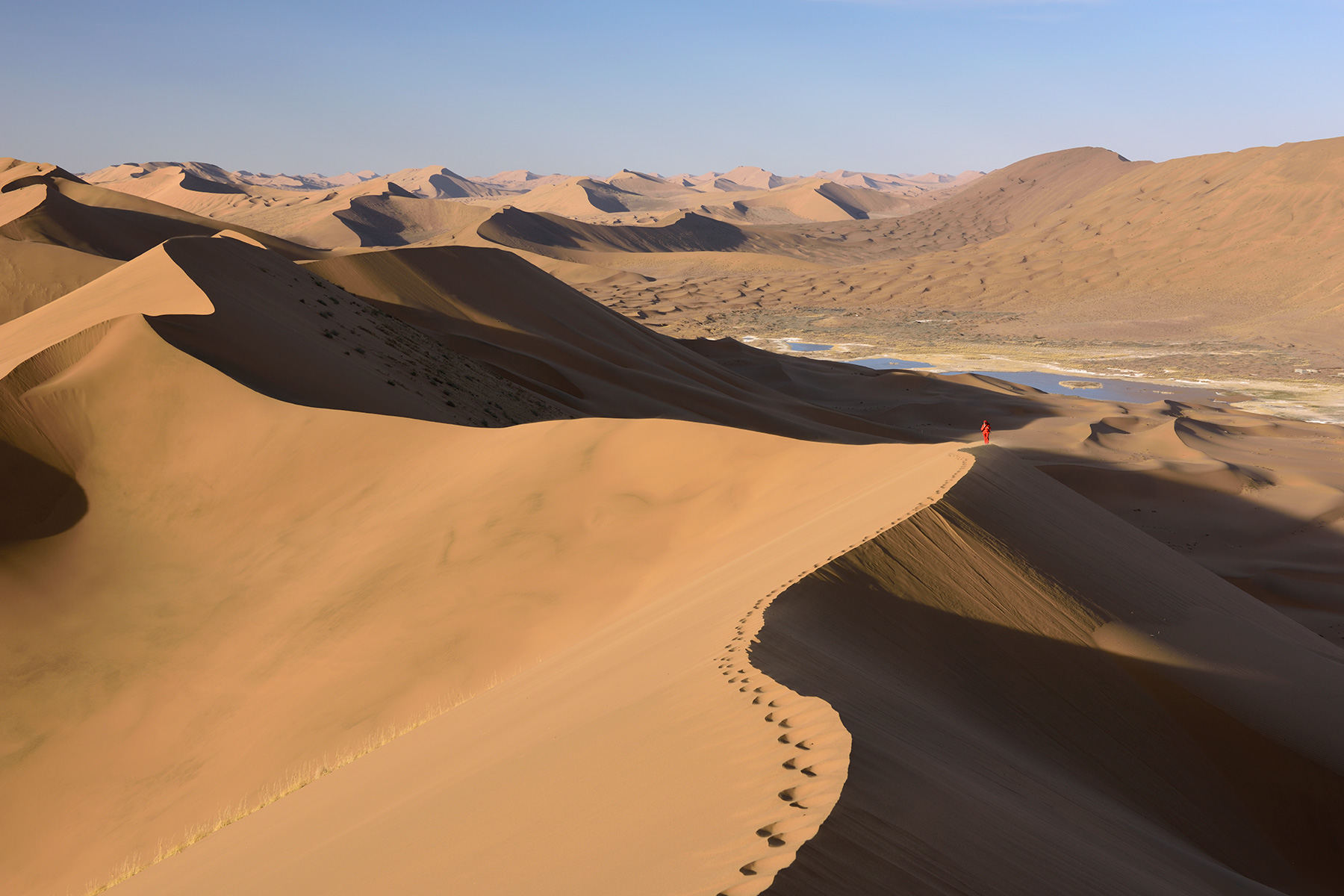 Désert du Badain Jaran (Chine, Mongolie Intérieure) - Vue générale d'un enchaînement de dunes avec randonneuse au loin sur la crête. Vallée avec lac salé à droite.