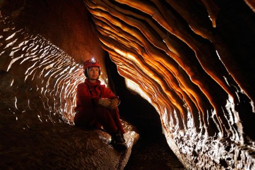 Grotte de l’Ascension (Hérault) - Spéléo assis dans petite galerie avec paroi couverte de draperies translucides (contre-jour)(Sp 10-0597)