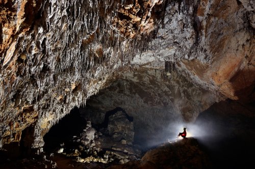 Dimnice Jama (Slovénie) - Spéléo assis de profil en contre-jour dans une grande salle regardant une paroi concrétionnée(SP-17-0497)