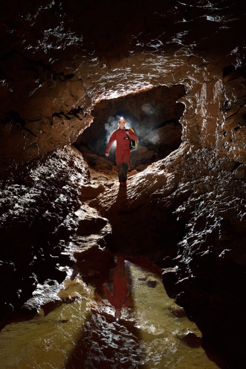 Grotte du sergent (Hérault) - Petite galerie rectangulaire  avec laisse d'eau. Réalisation d'une photo classique avec un contre-jour derrière le modèle et un flash de face dans un snoot. La buée autour du modèle (et la poussière secouée de la combinaison) matérialisent mieux le contre-jour. Un post traitement a été effectué sur l'eau, après l'avoir détourée, pour lui enlever du jaune.(SP-19-2083)