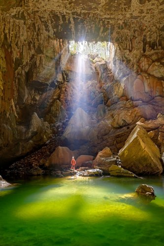 Lapa Terra Ronca (Goias, Brésil) - Rayons de soleil éclairant la rivière aux eaux vertes à partir d'un orifice (eau verte en premier plan et spéléo sur rocher à la base du gouffre). Format vertical.(SP-25-2020)
