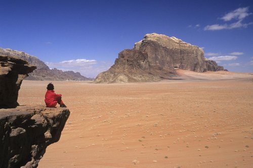  Désert du Wadi Rum. Larges étendues  de sable avec massifs de grès. Personnage assis sur un rocher surplombant. (VOY 06318)