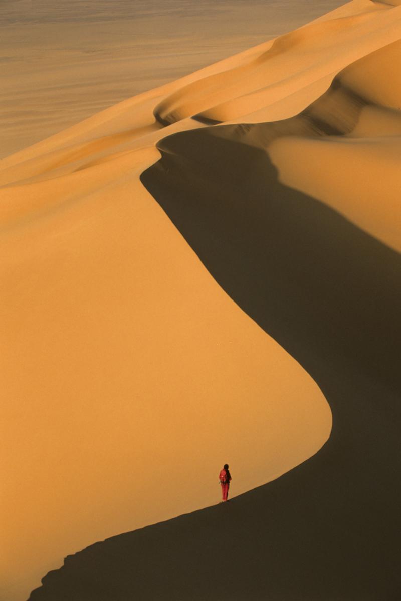 Femme (silhouette rouge) progressant sur la crête d'une dune au lever du soleil (Erg Wankasa, Désert de l’Akakus, Lybie)