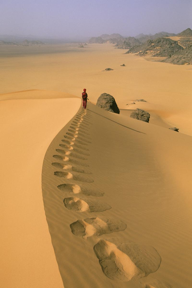 Femme de dos progressant sur la crête d'une dune avec traces de pas en premier plan (Wadi Aouis, Désert de l’Akakus, Lybie)